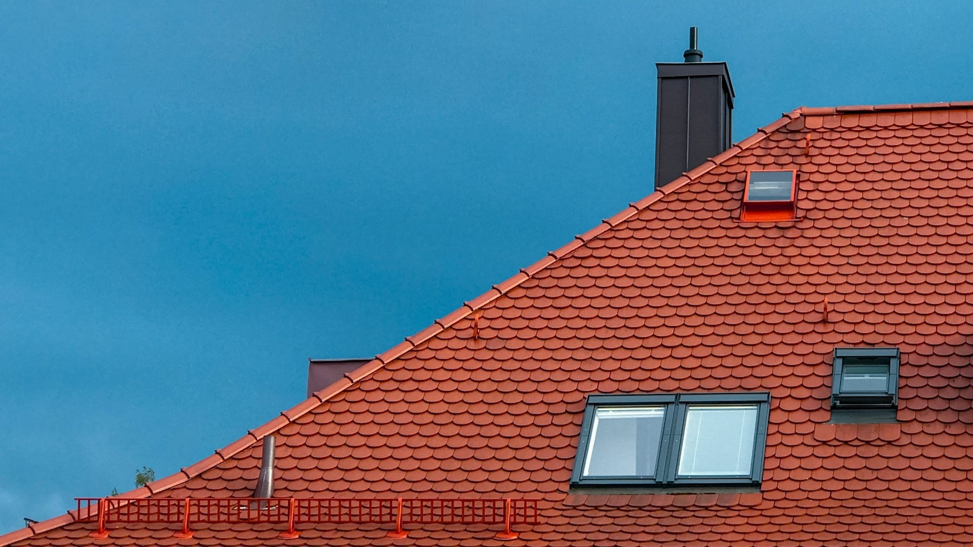 Rood pannendak met dakramen en schoorsteen onder een heldere blauwe lucht, passend bij het thema hoe lang gaat een dak mee