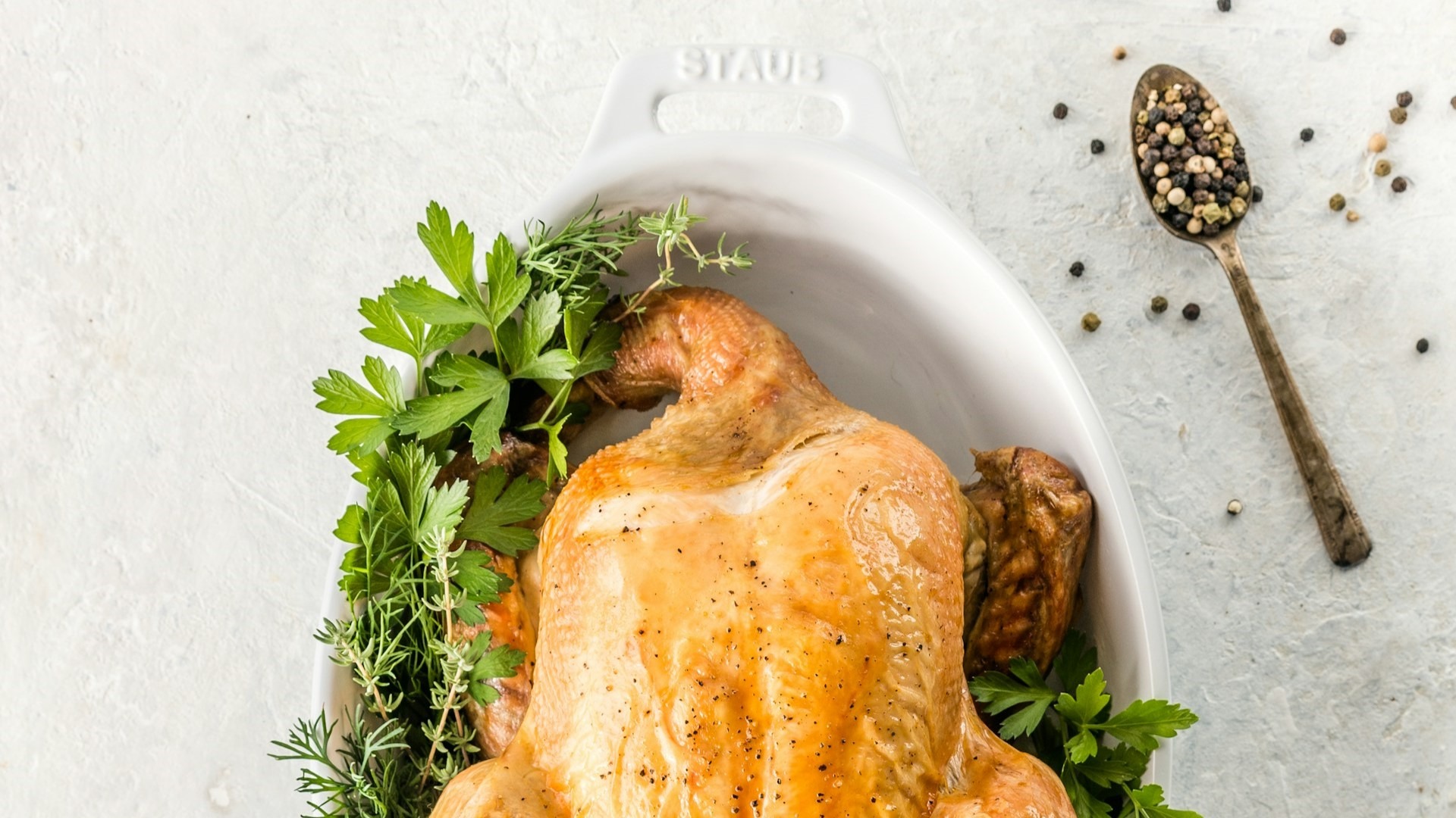 Hele kip braden met verse kruiden in een ovenschaal, omringd door peterselie, tijm en peperkorrels