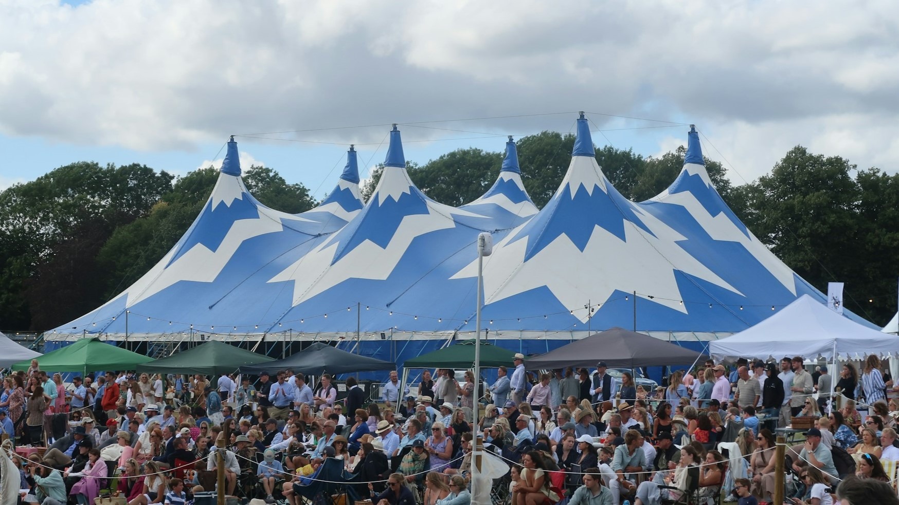 Grote blauw-witte festival tent met pieken, omringd door een menigte bezoekers op een buitenfestival