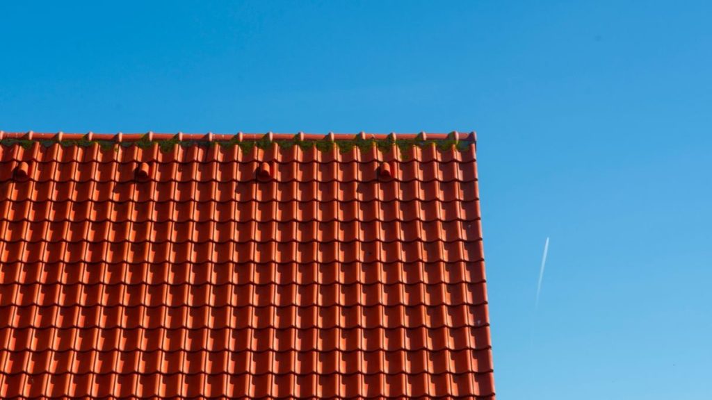 Rode dakpannen op puntdak met wat mosgroei, tegen een strakblauwe lucht met een condensspoor